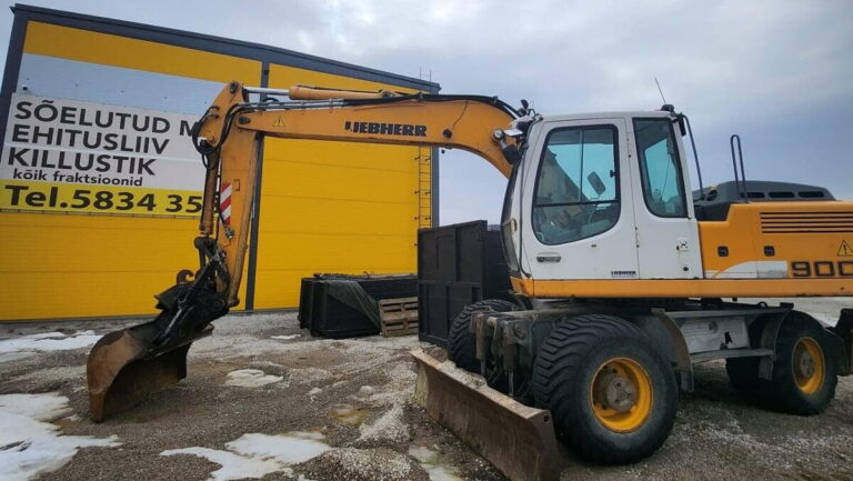 Kollane Liebherr ekskavaator killustikuplatsi ees, taustal kollane hoone materjalimüügi reklaamiga.